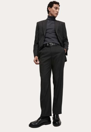 Dunkelgrauer, maßgeschneiderter Anzug mit schmaler Passform, kombiniert mit einem schwarzen Rollkragenpullover. Schwarzer Gürtel und polierte elegante Schuhe vervollständigen den Look.