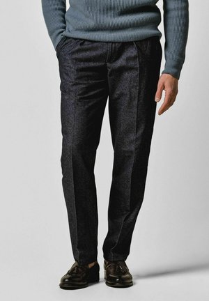 Man draagt donkergrijze nette broek, bruine loafers met kwastjes en een blauw geribde trui met één hand in de zak tegen een effen achtergrond.