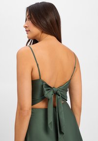 Groene satijnen top met een lage rug, dunne bandjes en een grote strikdetail aan de achterkant. De stof heeft een gladde, glanzende textuur.