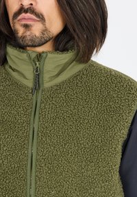 Vihreä fleece-vesti, jossa on teksturoitu, pörröinen ulkopinta ja sileä sisäkaulus; varustettu etuvetoketjulla ja korkealla kaulussuunnittelulla.