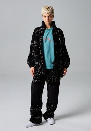 Black patterned fleece jacket and pants with wavy line design, paired with a light blue t-shirt featuring “GRMY” in pink. White sneakers.