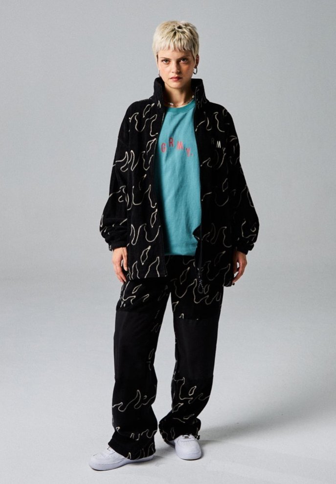Zwarte fleecejack en -broek met een golfpatroon, gecombineerd met een lichtblauw T-shirt met "GRMY" in het roze. Witte sneakers.