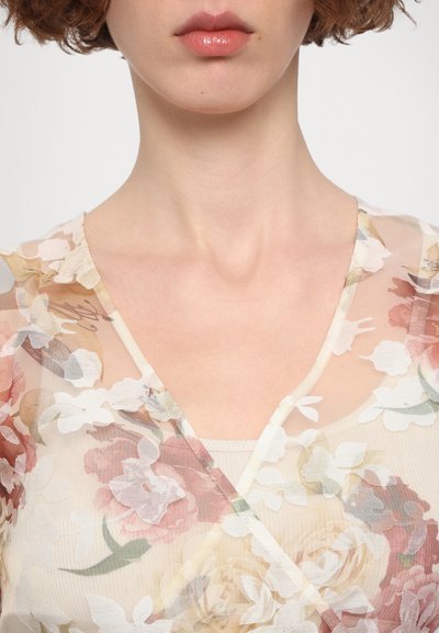 Florale doorschijnende top van lichte stof, met een V-hals ontwerp en roze en witte bloemenpatronen op een beige achtergrond. Subtiele textuurdetails.
