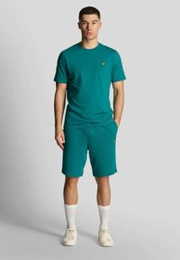 T-shirt unisex in cotone color verde petrolio e pantaloncini abbinati, con un piccolo logo giallo. Il modello indossa calzini bianchi alti e sneakers bianche.
