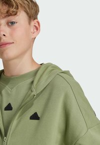Sudadera verde claro hecha de una tela suave, con cremallera en la parte delantera y dos acentos del logo negro de Adidas cerca del cuello.