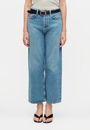 Žena nosi plave traperice visokog struka širokih nogavica s crnim remenom i crnim sandalama otvorenih prstiju, stoji ispred jednostavne pozadine.