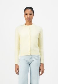 Cardigan giallo a maglia con maniche lunghe, collo rotondo e chiusura con bottoni. Tessuto morbido, forma aderente, abbinato a jeans blu chiaro.