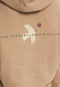 Béžová mikina s kapucí se stylizovaným logem ptáka a nápisem „Středozemní mořská fauna“ na zádech.