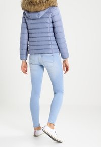 Ljusblå puffjacka med foder avpälsad huva, horisontellt quiltad design, i kombination med ljusblå jeans och vita sneakers.