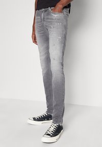 Vaqueros de mezclilla gris claro con detalles desgastados, corte ajustado y un suave desvanecimiento. Combinados con zapatillas de lona negras y una camiseta negra.