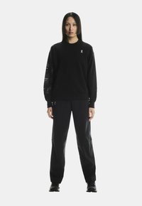 Fekete pulcsi, egy nyomott dizájnnal az egyik ujjon, fekete nadrággal párosítva. Az öltözet laza szabású és puha tapintású.