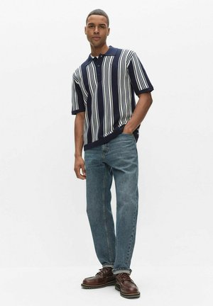 Mężczyzna stojący z jedną ręką w kieszeni, ubrany w granatowo-białą, pionowo prążkowaną koszulkę polo z krótkim rękawem, niebieskie dżinsy i brązowe skórzane buty.