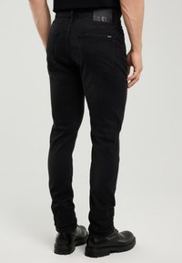 Férfi fekete slim fit farmernadrágban  és fekete bőr cipőben, háttal a kamerának áll egy egyszerű, világos háttér előtt.