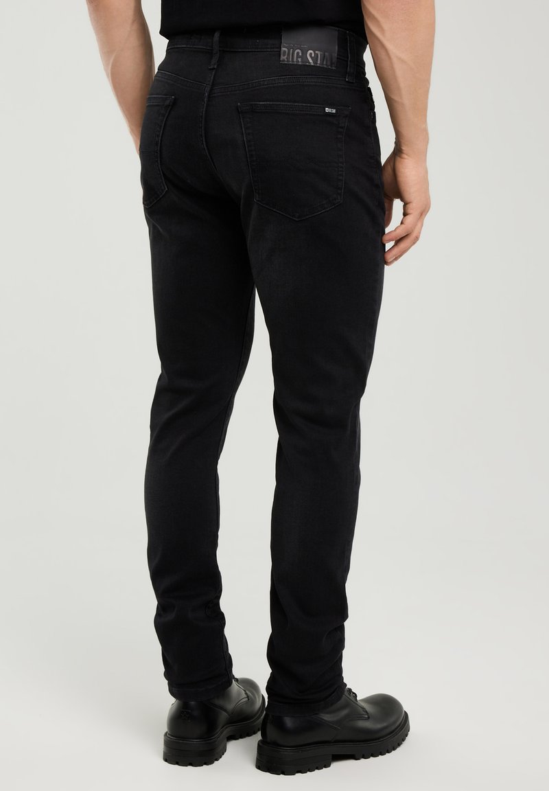 Férfi fekete slim fit farmernadrágban és fekete bőr cipőben, háttal a kamerának áll egy egyszerű, világos háttér előtt.