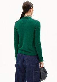 Vrouw met donker haar in een paardenstaart, die een groene sweater en blauwe spijkerbroek draagt, terwijl ze een grijze pet vasthoudt, met de rug naar de camera gekeerd.