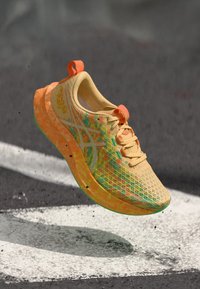 Light orange and yellow running shoe with speckled sole floating above asphalt with white painted lines.