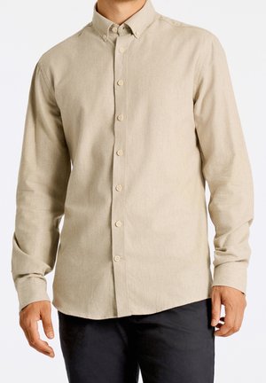 Mand iført en beige skjorte med knapper og lange ærmer med krave, kombineret med sorte bukser, stående foran en hvid baggrund.