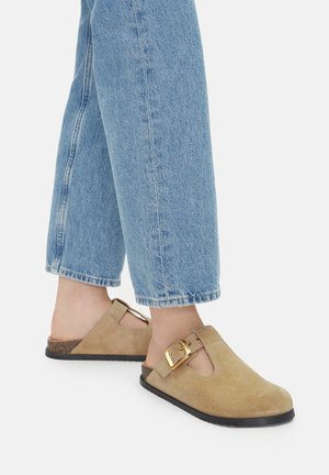 Béžové semovité dřeváky s otevřenou zadní částí, dekorativní přezkou a černou gumovou podrážkou. Nosené s světle modrými kalhotami s uvolněným střihem a ohrnutým lemem.