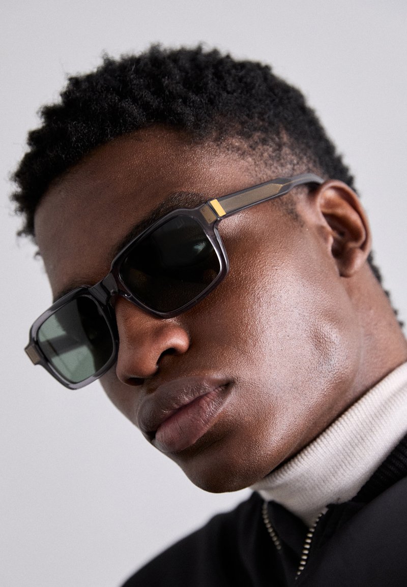 Γυαλιά ηλίου με σκούρους φακούς, ορθογώνιο σκελετό και υφασμάτινες βραχίονες με χρυσή λεπτομέρεια. Φοριούνται με ανοιχτόχρωμο ζιβάγκο και σκούρο σακάκι.