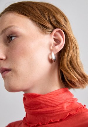 Silver teardrop earring with a polished finish, worn by a person in a red ribbed, high-neck top. Hair is styled in loose waves.