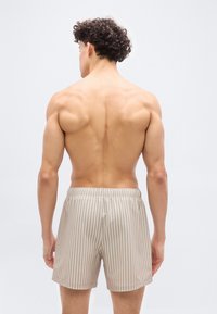 Νεαρός με σγουρά μαλλιά, γυρισμένος ανάποδα, φοράει μπεζ και λευκά κάθετα ριγέ σορτς σε απλό φωτεινό φόντο.