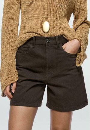 Personne portant un pull en tricot marron et un short marron foncé, une main dans la poche et un collier pendentif ovale doré.