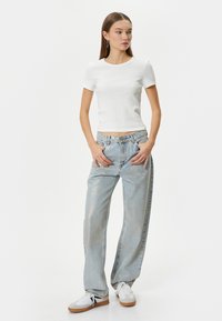 Hvid tætsiddende t-shirt, lyseblå baggy jeans med falmet tekstur og hvide sneakers med sorte og brune accenter. Modellen poserer med hænderne på hofterne.