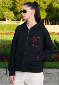 Jeune femme avec une longue queue de cheval noire porte un sweat à capuche noir zippé avec un motif de cœur rouge et un pantalon blanc, debout à l'extérieur dans un jardin.