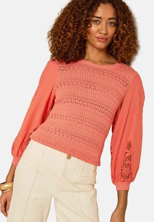 Kvinde med krøllet hår iført en koralfarvet bluse med hæklet mønster og lange ærmer samt højtaljede cremefarvede bukser med flettede lommedetaljer.