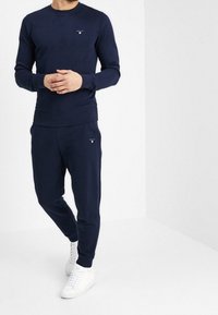 Man som bär en marinblå sweatshirt och matchande joggers, ihop med vita sneakers, står mot en enkel ljus bakgrund.