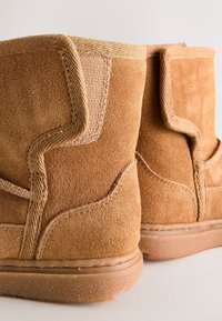 Paire de bottes en daim beige avec semelles en caoutchouc texturées et coutures renforcées autour du haut et des côtés, visibles de dos.