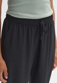 Schwarze Sporthosen mit einem Kordelzugbund, ausgestattet mit seitlichen Taschen und einem glatten, leichten Stoff. Model trägt ein hellgrünes Oberteil.