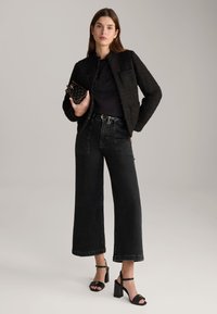 Zwarte textuurjack met een ronde halslijn, gecombineerd met losse zwarte denim jeans en zwarte hoge hakken sandaaltjes. Model houdt een kleine tas vast.