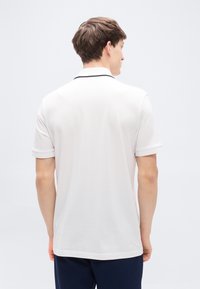 Πολύ κοντομάνικο πόλο πουκάμισο σε ανοιχτό μπεζ με ριγωτή υφή και μαύρο περιθώριο στο γιακά. Η πλάτη έχει ευθεία τελείωμα και διακριτικό ράψιμο.