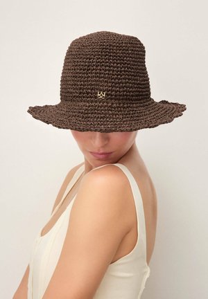 Femme portant un chapeau bob brun en crochet et un haut crème sans manches, regardant vers le bas sur un fond uni.