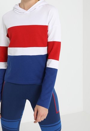 Persona que lleva una sudadera con capucha de bloques de color con rayas blancas, rojas y azules y mallas deportivas azules a juego con detalles en rojo y azul.