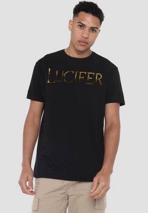 Young man with short curly hair wearing a black T-shirt with shiny gold "LUCIFER" text and beige cargo shorts, standing against a gray background.