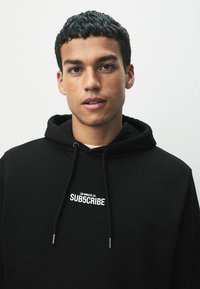 Juoda hoodie su didele priekiu kišene, su raišteliu ant gaubto ir balta siuvinėta užrašais "SUBSCRIBE" ir "LOS ANGELES, CA." Lygus paviršius.