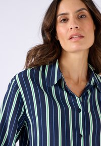 Chemise marine à rayures verticales vertes et bleues. Dotée d'un col classique et de boutons, fabriquée dans un tissu lisse avec un léger brillant.