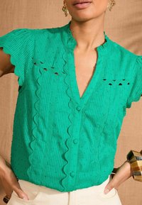Green blouse with short ruffled sleeves, vertical stripes, textured bobble pattern, and scalloped edges. Features a V-neck and button closure.
