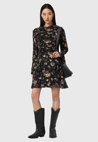 Vestido negro con estampado floral, mangas largas, corsé con volantes y falda de capas, combinado con botas vaqueras negras y un bolso negro.