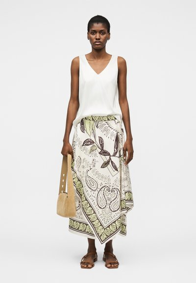 Γυναίκα που φοράει λευκό τοπ χωρίς μανίκια, midi φούστα με φύλλα και σχέδιο παίζλι, καφέ σανδάλια και κρατάει χειροποίητη τσάντα από καφέ σουέτ.