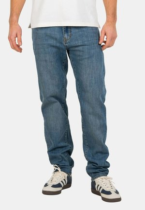 Blaue Jeans aus Denim mit geradem Bein und sichtbaren Nähten; getragen mit weißen und marineblauen Sneakers. Verfügt über normale Taschen und einen Reißverschluss.