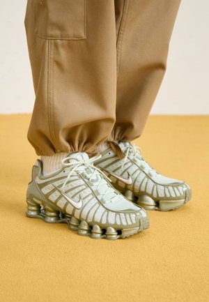 Jasne buty do biegania Nike z amortyzującą podeszwą sprężynową, noszone z beżowymi spodniami dresowymi na musztardowej podłodze.