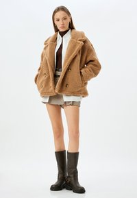 Giacca in pile marrone con ampi rever, design doppiopetto e tasche laterali. Indossata sopra una camicia bianca e un top a coste marrone, abbinata a pantaloni corti.
