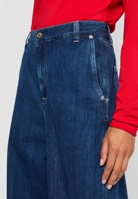 En person som har på seg mørkeblå denimjeans og en langermet rød skjorte, med den ene hånden hvilende ved siden av seg.