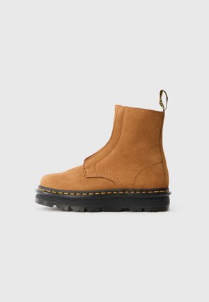 Bottine en cuir beige avec une semelle épaisse noire, des coutures jaunes et une languette à l'arrière sur fond gris.