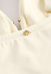 Cream bikini top featuring textured fabric, adjustable straps, and a gold-tone circular clasp detail at the center.