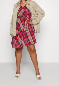 Žena oblečená v béžové prošívané bundě přes červené, zelené a žluté kostkované šaty s zavázaným pasem, doplněná o bílé otevřené sandály na podpatku.