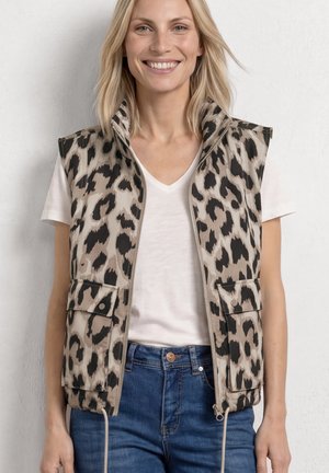 Femme souriante portant un gilet zippé beige et noir à imprimé léopard par-dessus un T-shirt blanc à col en V et un jean bleu, debout devant un mur clair.